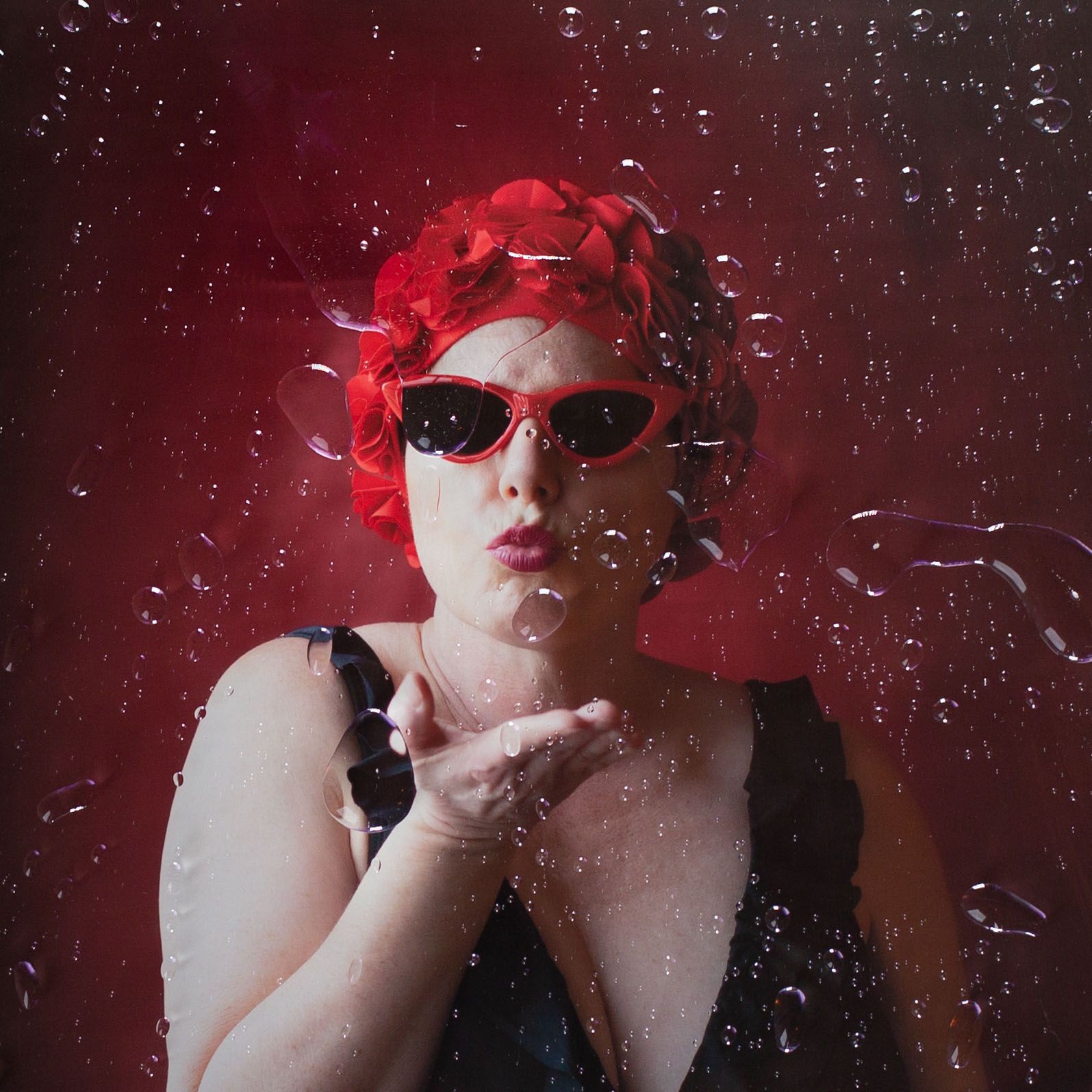 Woman with red hair and sunglasses blowing glitter against a dark red background