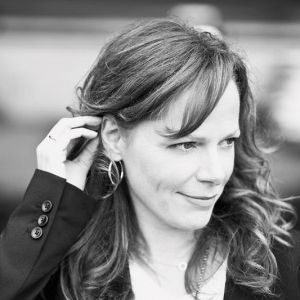 Portrait of a person adjusting their hair, wearing hoop earrings and a dark coat, in black and white.