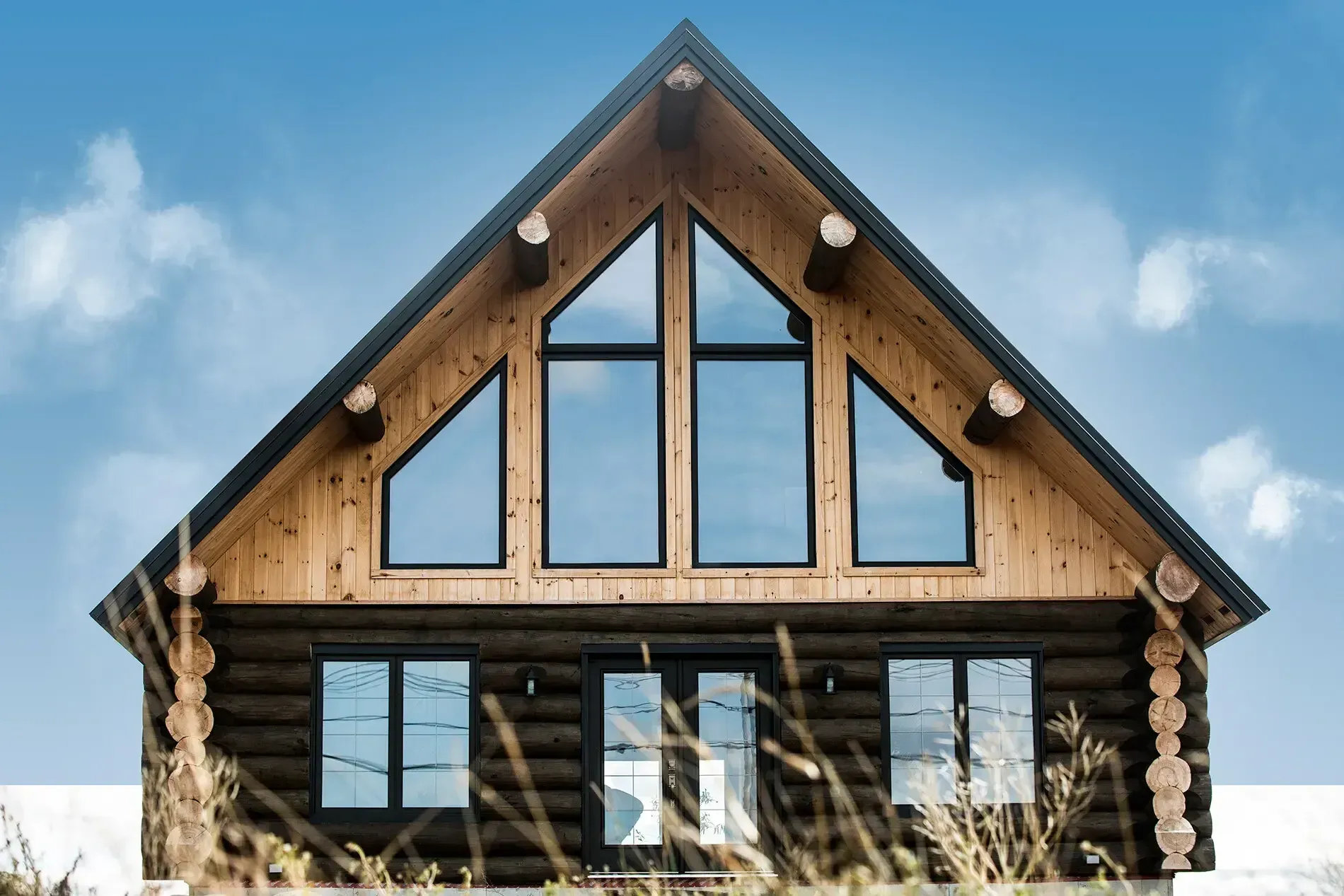 Cabane en rondins avec de grandes fenêtres triangulaires, rondins bruns, boiseries noires, ciel bleu en arrière-plan.