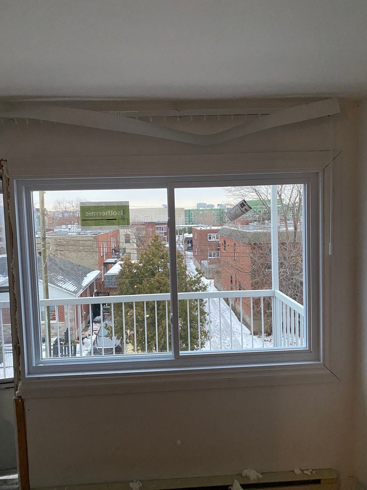 Vue depuis une fenêtre donnant sur un balcon enneigé, des immeubles et des arbres. Cadre de fenêtre blanc, ciel nuageux.