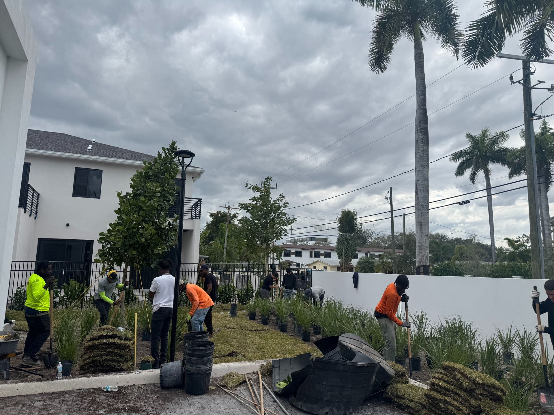 A crew of workers in bright safety vests plants vegetation along a white wall next to a house on a cloudy day.