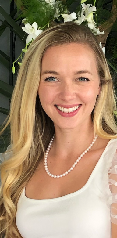 Smiling person with long blonde hair and a pearl necklace in front of a white floral backdrop.