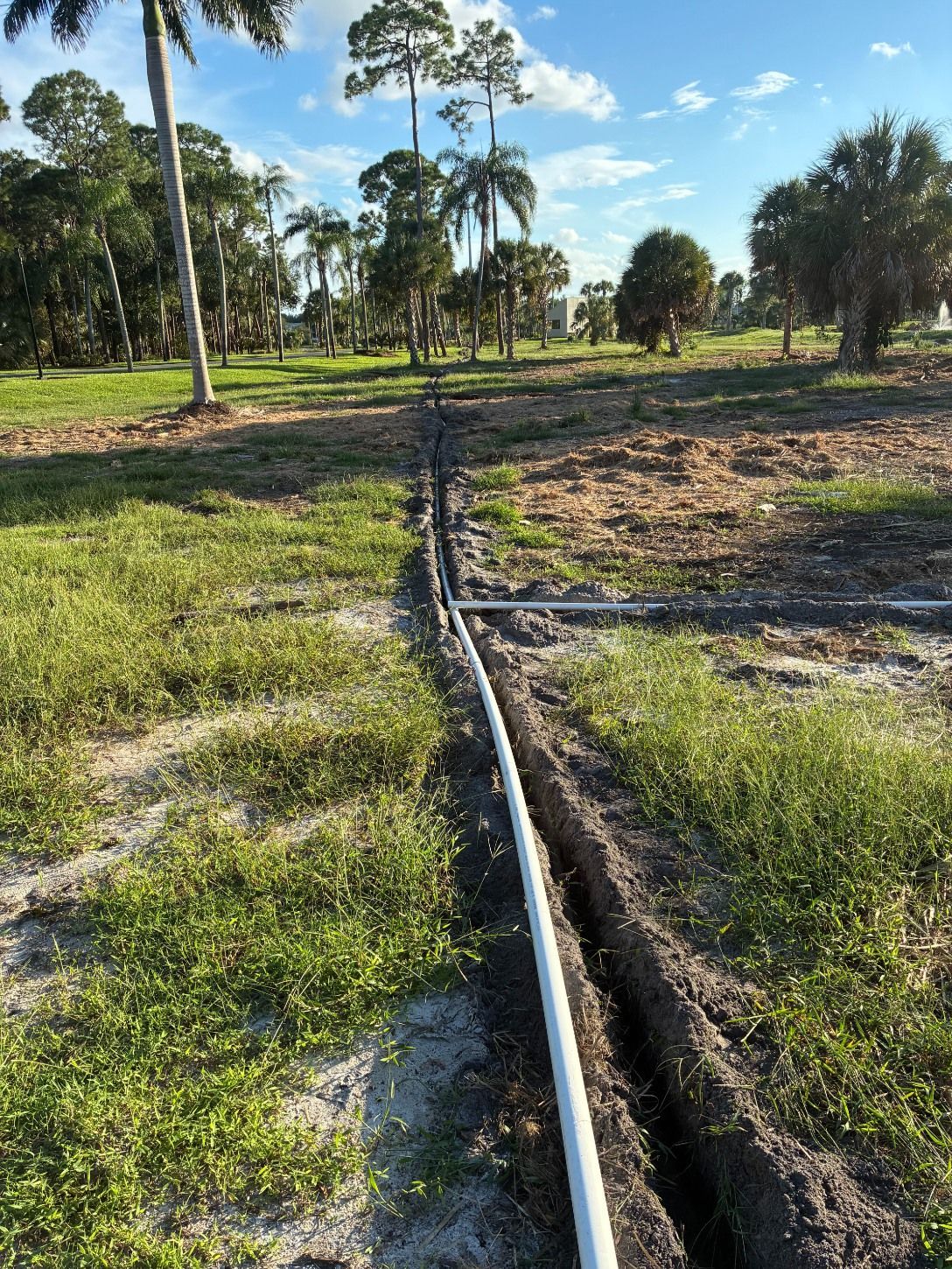 White PVC pipes lay in a narrow, unpaved trench cut through a grassy field with trees in the background.