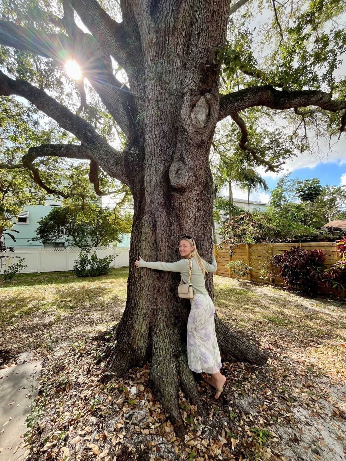 A person in a light top and floral skirt hugs a large, textured tree trunk in a sunlit yard.