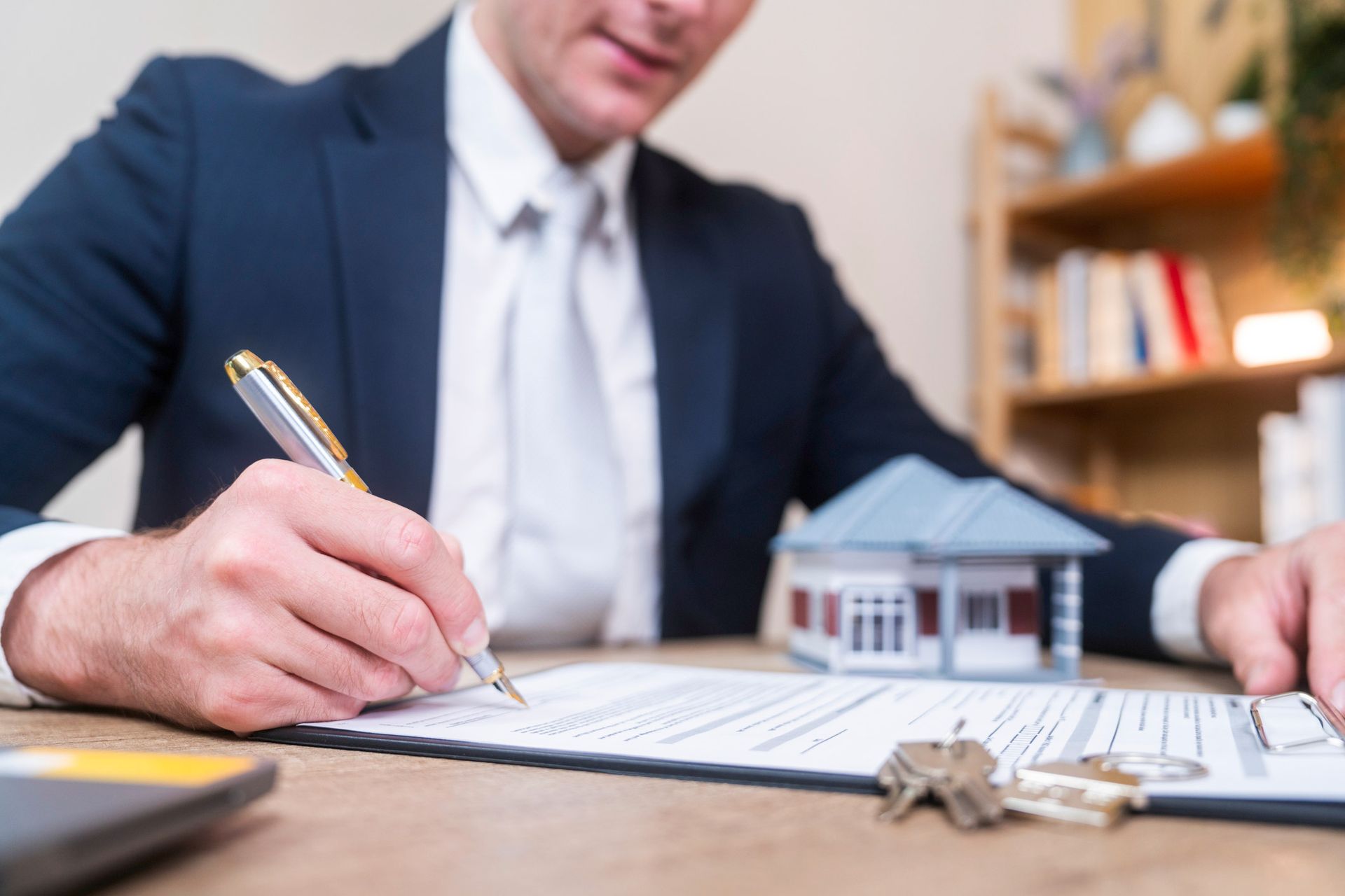 Person signing a real estate contract with house model and keys on the table.