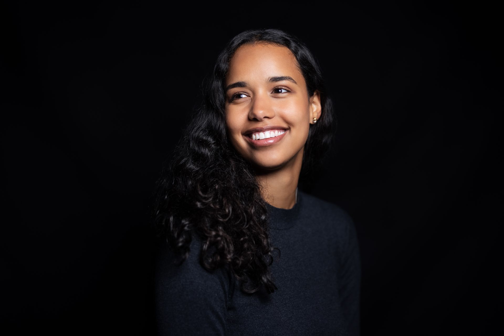 A woman with curly hair is smiling in front of a black background.