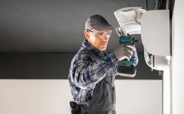 Technician in safety glasses and cap repairs a wall-mounted device with a power drill.