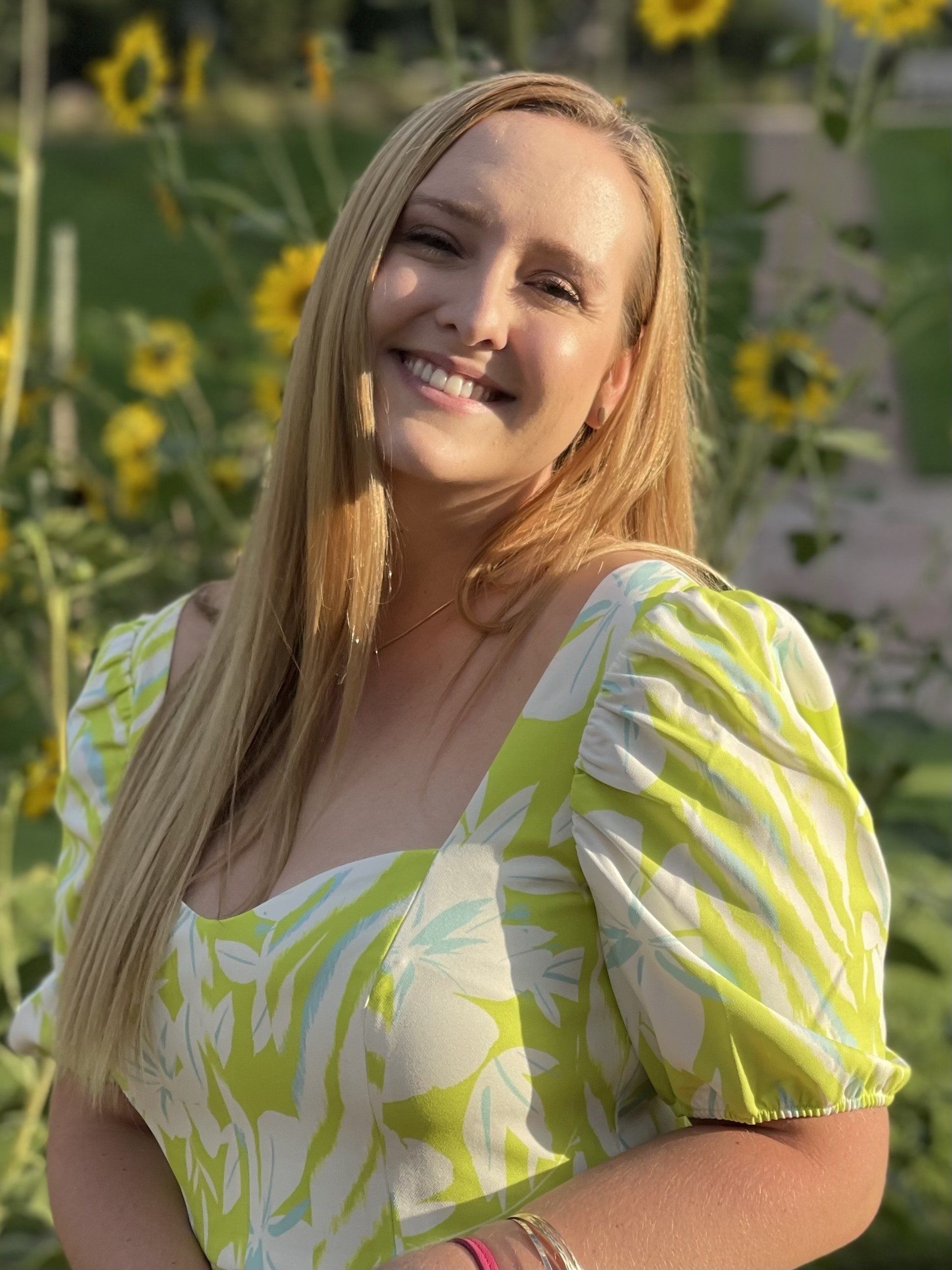 A woman in a green and white dress is smiling in front of a field of sunflowers.