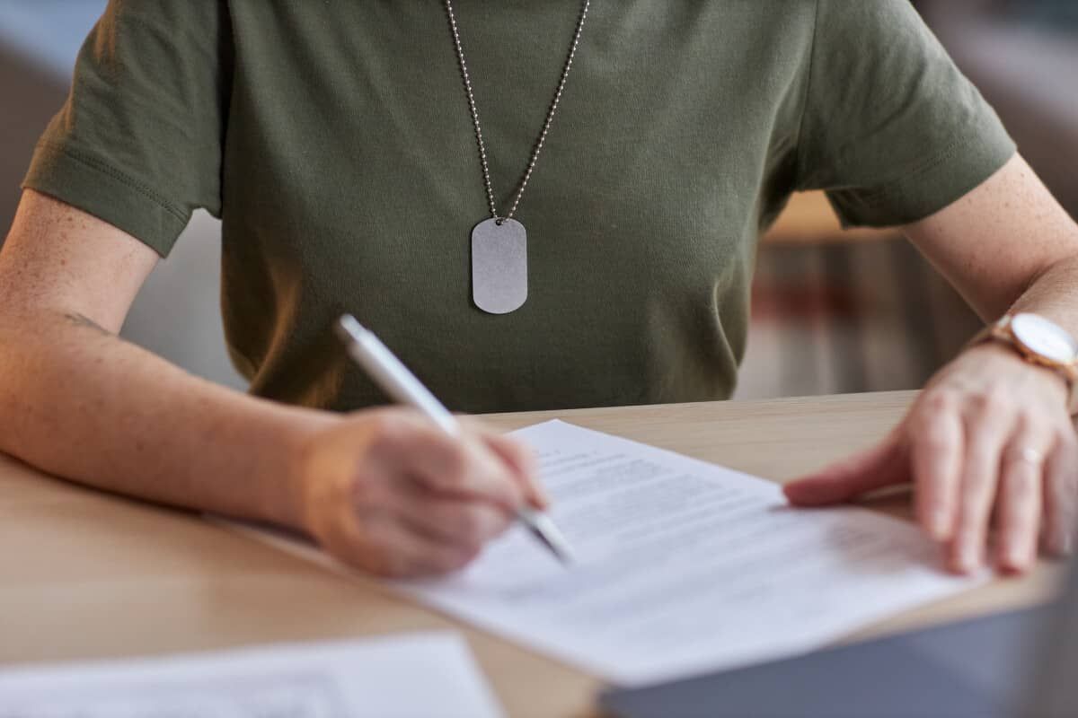 Person signing a document at a desk, wearing a green shirt and dog tag necklace