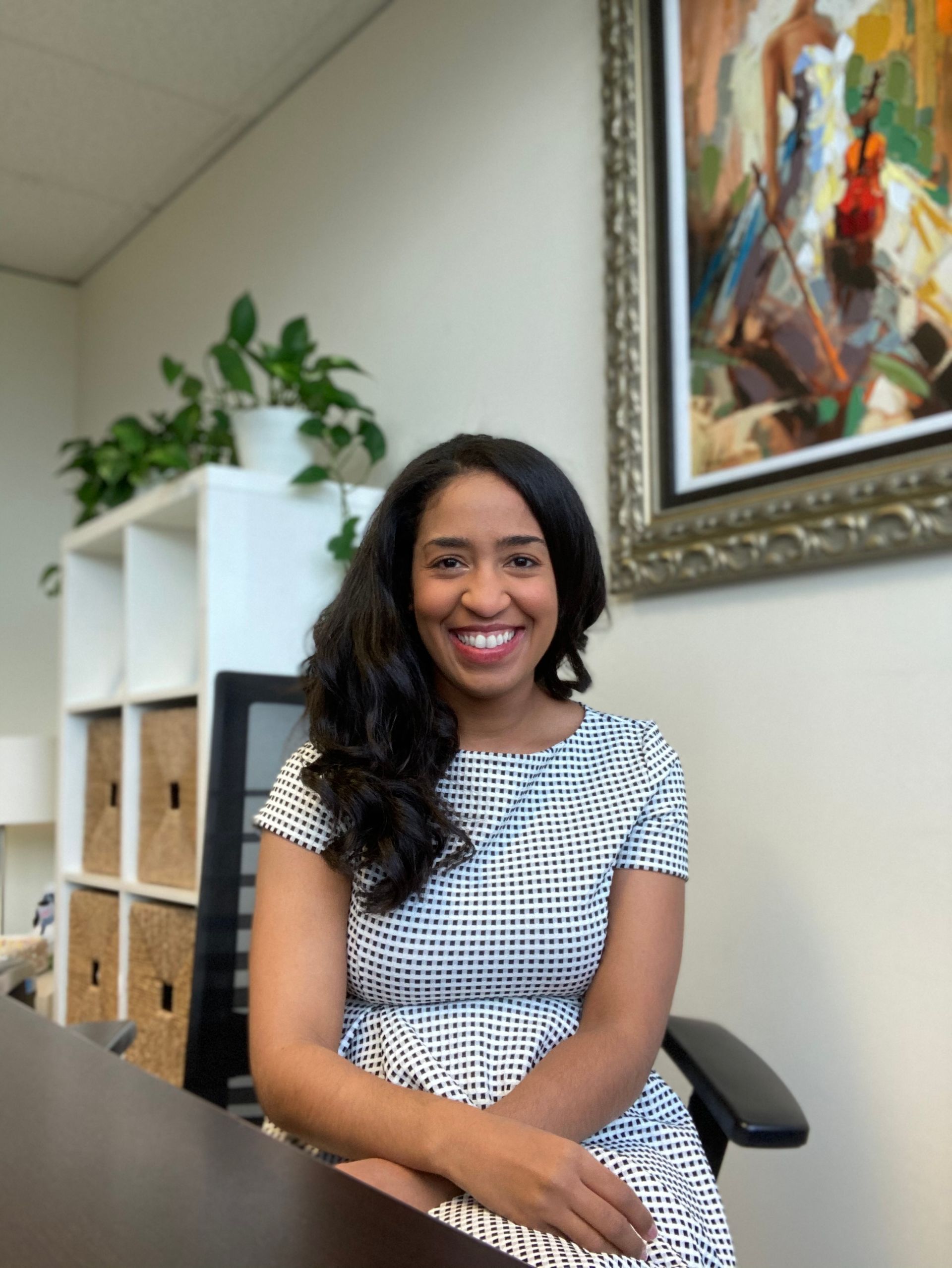Woman smiling in an office chair beside shelves, a plant, and colorful wall art.