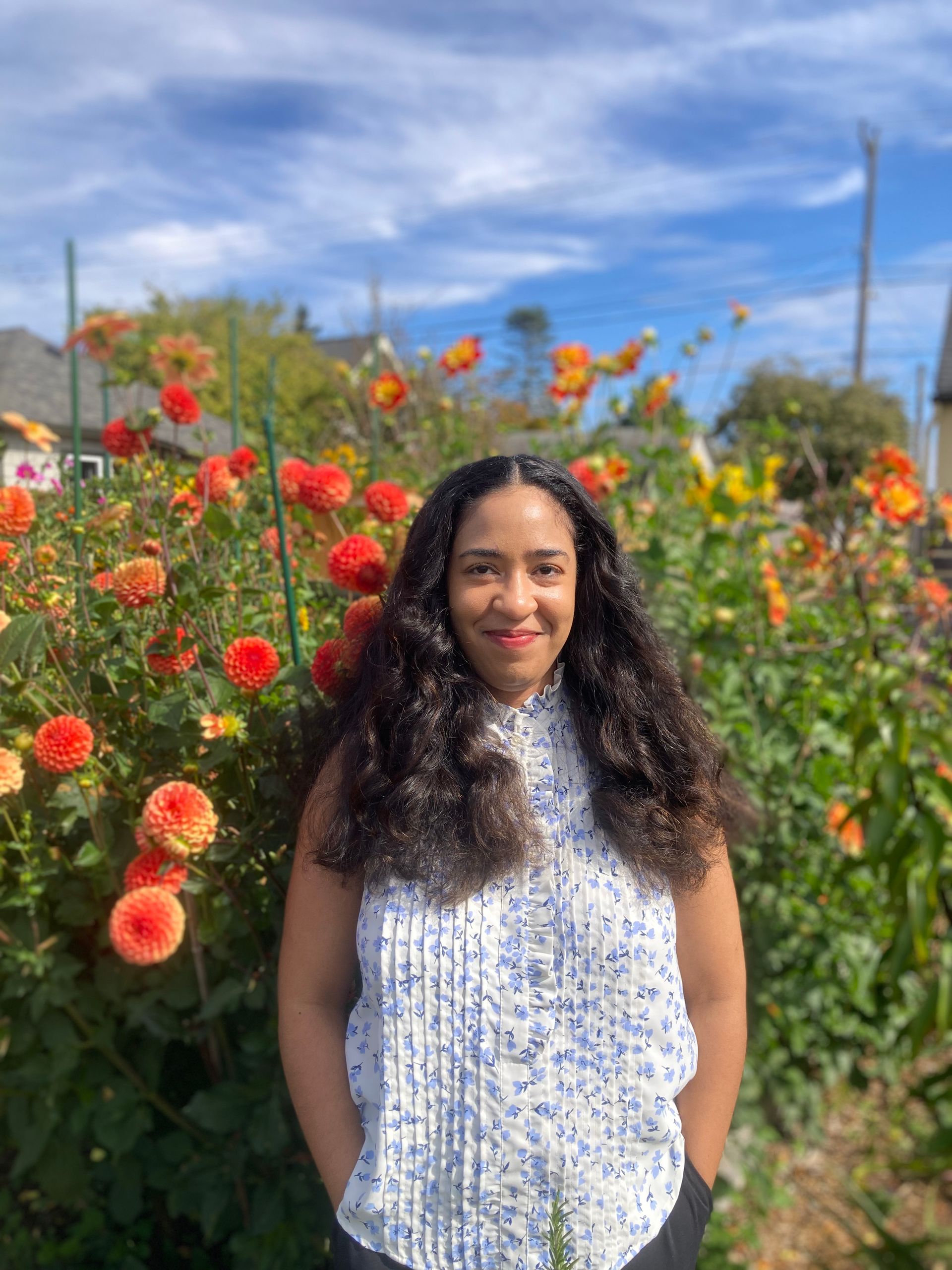 Woman smiling outdoors, wearing a blue patterned shirt, near a tree and greenery.