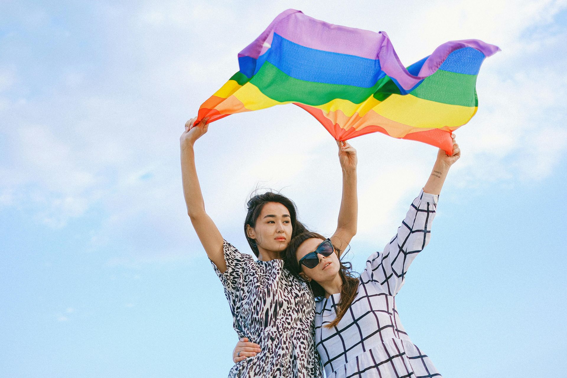 Two people holding a rainbow Pride flag overhead against a blue sky