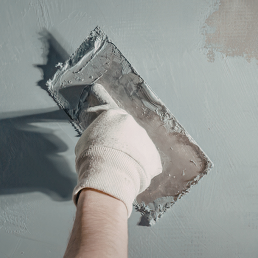 Gloved hand smoothing gray plaster on a wall with a trowel
