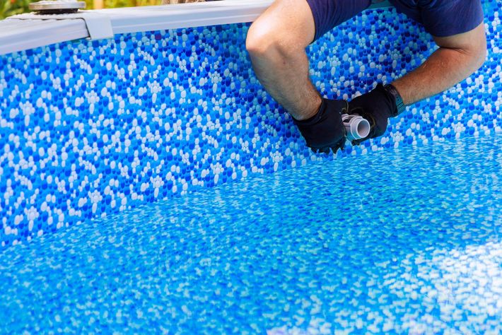 Una persona inginocchiata in una piscina rivestita di mosaici blu tiene in mano un oggetto nero vicino all'acqua.