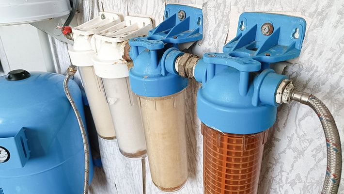 Tre alloggiamenti blu per filtri dell'acqua con cartucce bianche e marroni montati a parete e collegati tramite tubi flessibili.