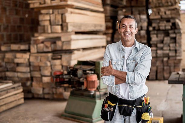 Smiling carpenter in a woodshop with stacked lumber and tools on his belt