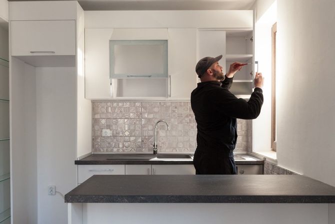 Man in black shirt adjusting a wall cabinet in a modern white kitchen