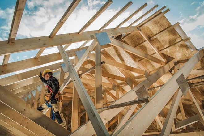 Construction worker in a wooden roof frame, wearing a hard hat and harness under blue sky.
