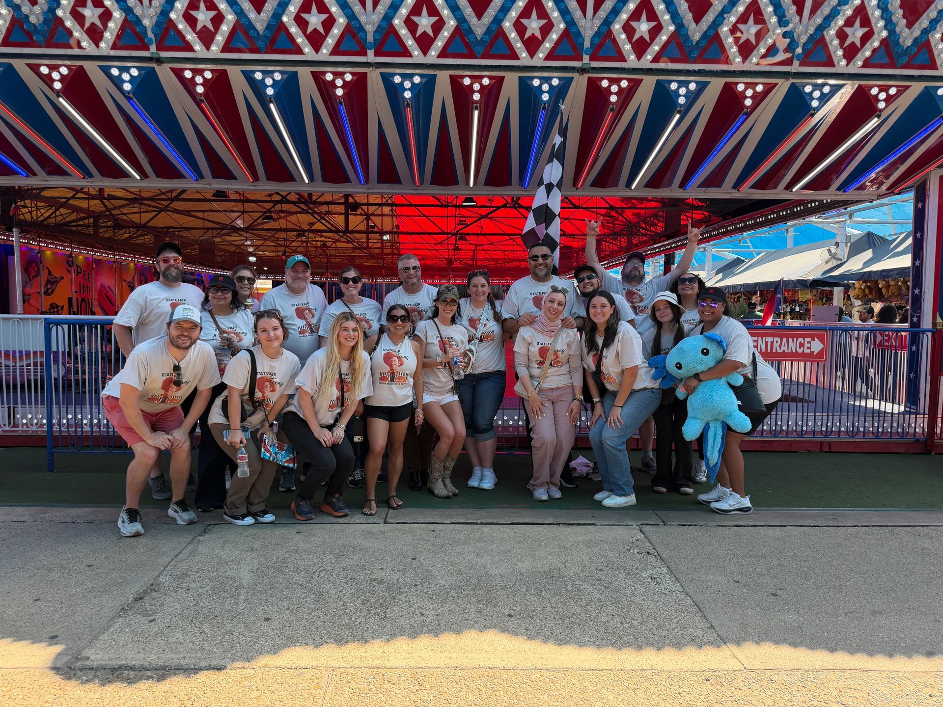 Group of people posing in front of a red-and-blue carnival ride, with one person holding a large blue stuffed animal at the State Fair of Texas
