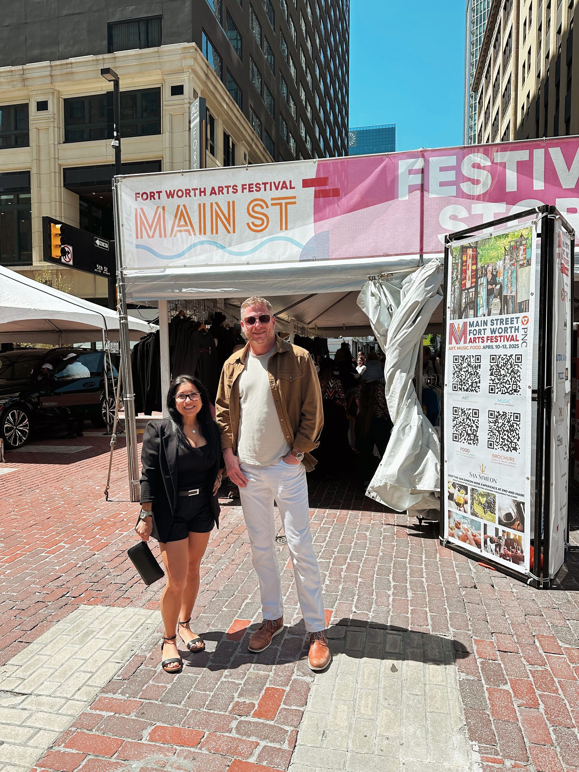 Two people posing at a Main St festival booth with tents, banners, and downtown buildings in the background.