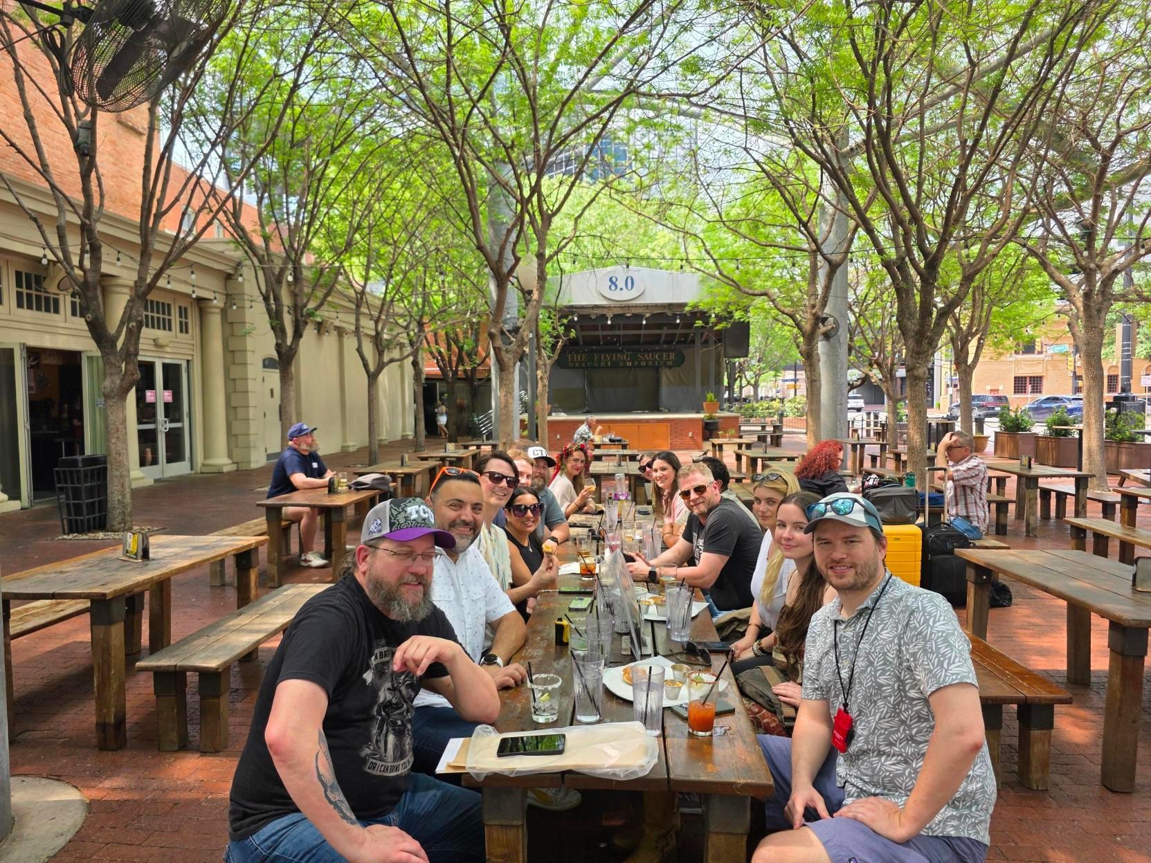 Group seated at an outdoor café patio under leafy trees, sharing drinks and a meal
