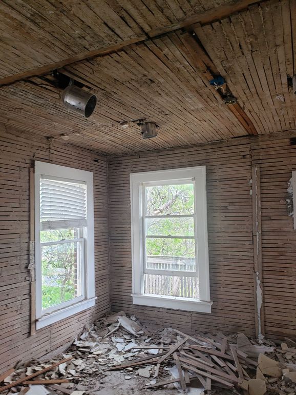 An interior room undergoing renovation with exposed wood lath on walls and ceiling, two windows, and debris on the floor.