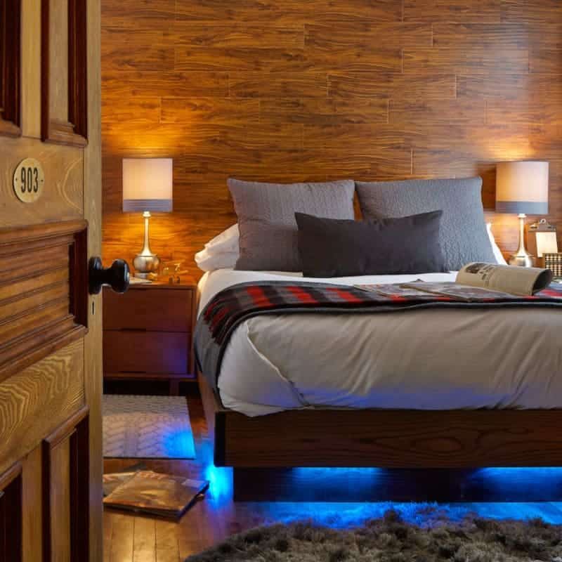 A bedroom with a bed , nightstand , lamps and a wooden wall.
