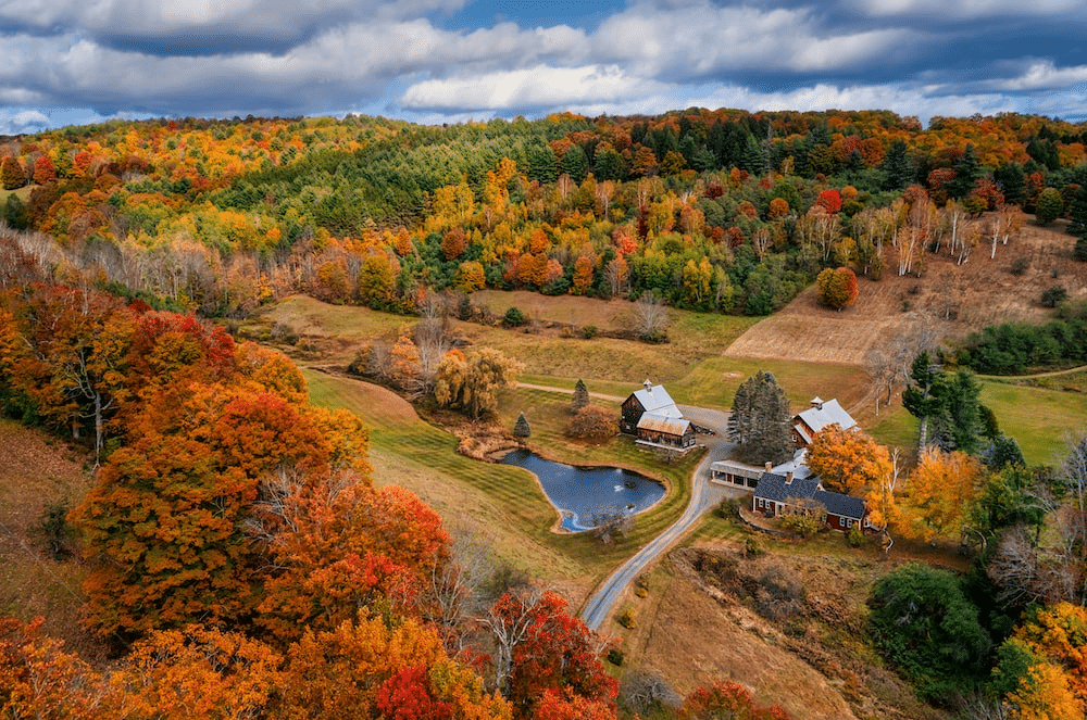 Rolling hills ablaze with fall colors overlook a farmhouse with a pond, New England.