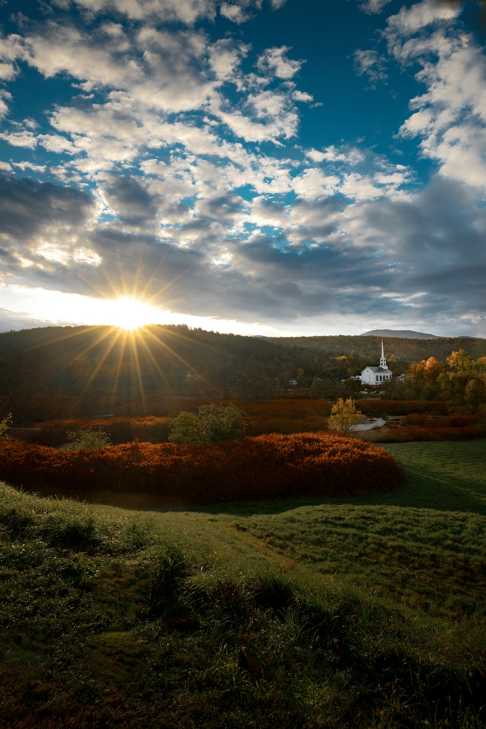 Sunset over a valley with a white church; vibrant sky, red and green foliage.