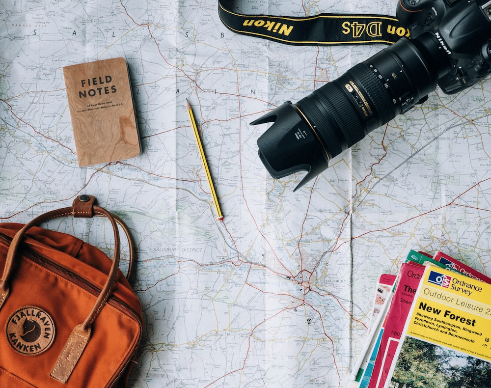 Overhead view: orange bag, camera, map, notepad, pencil, travel brochures, all on a map.