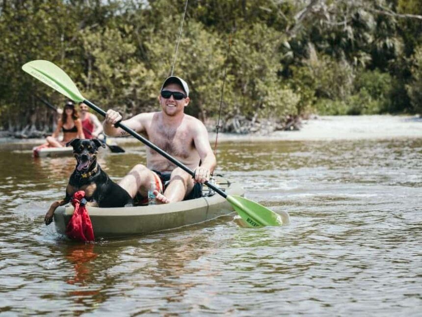 Man kayaking with dog, shirtless, on sunny water, with another person in the background.