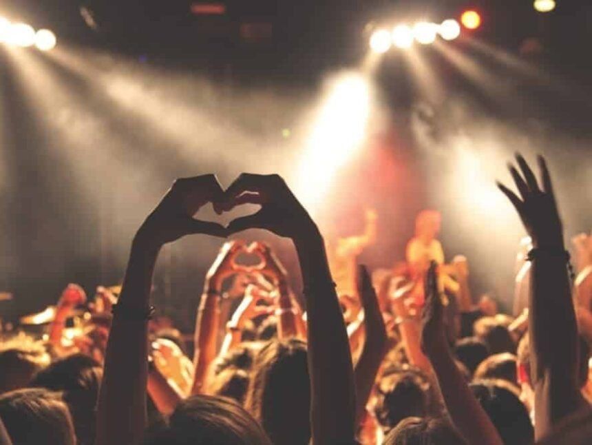 Concert crowd, many arms raised, one making a heart shape, illuminated by stage lights.