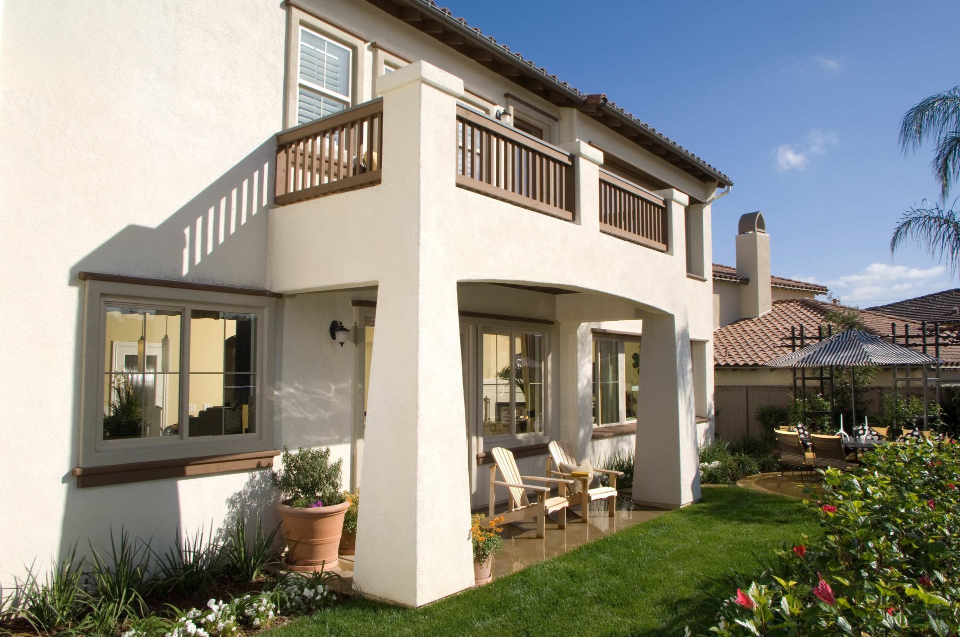 Two-story stucco home with balconies, patio, and a sunny backyard with grass and flowers. Two-story stucco home with balconies, patio, and a sunny backyard with grass and flowers.
