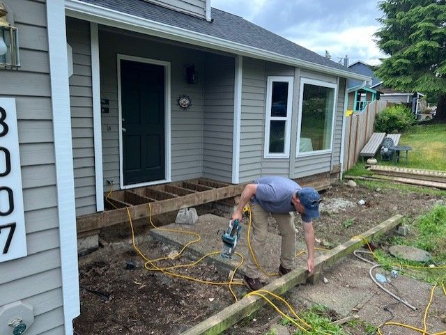 Man using a drill to build a deck in front of a house. Gray siding, green lawn, and yellow cords are visible.