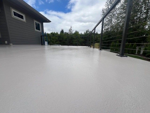 Gray deck with black railing, overlooking trees and blue sky. House siding visible.