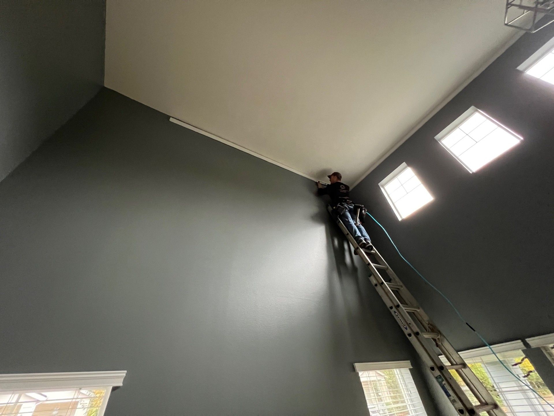 Person on tall ladder painting the trim of a high, gray wall and white ceiling.