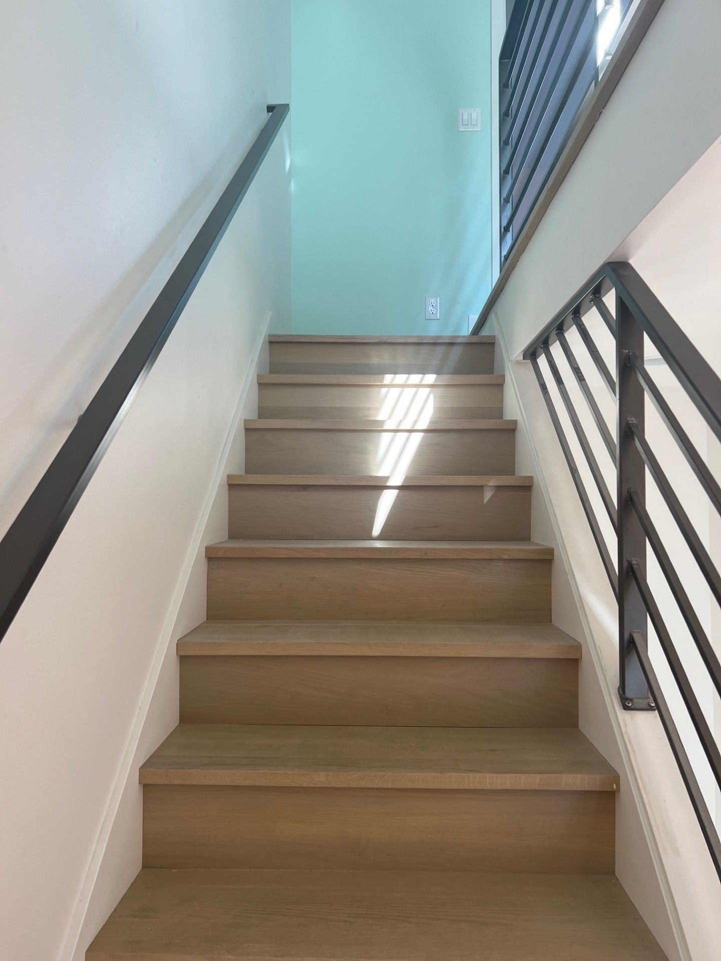 Staircase ascending to light blue wall, with wooden steps and black handrails.
