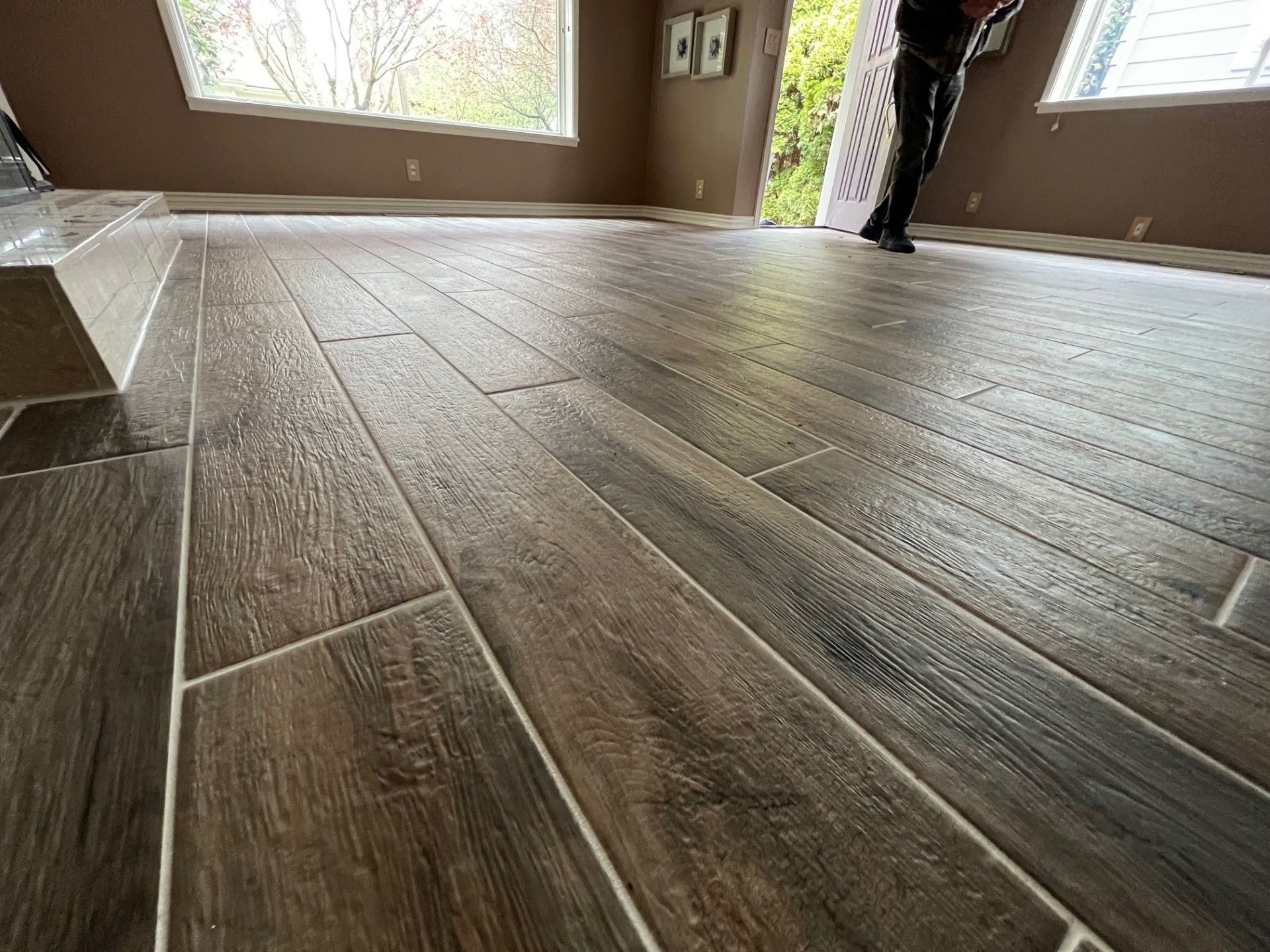 Close-up of a room with newly installed, wood-look tile flooring. Person in background. Brown walls and trim.