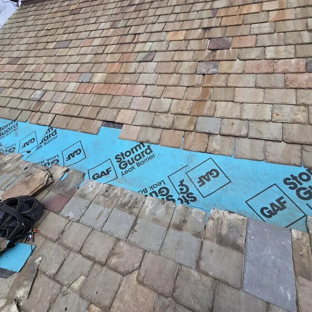 A roof under renovation, showing a blue GAF StormGuard underlayment strip between sections of rectangular brown shingles.