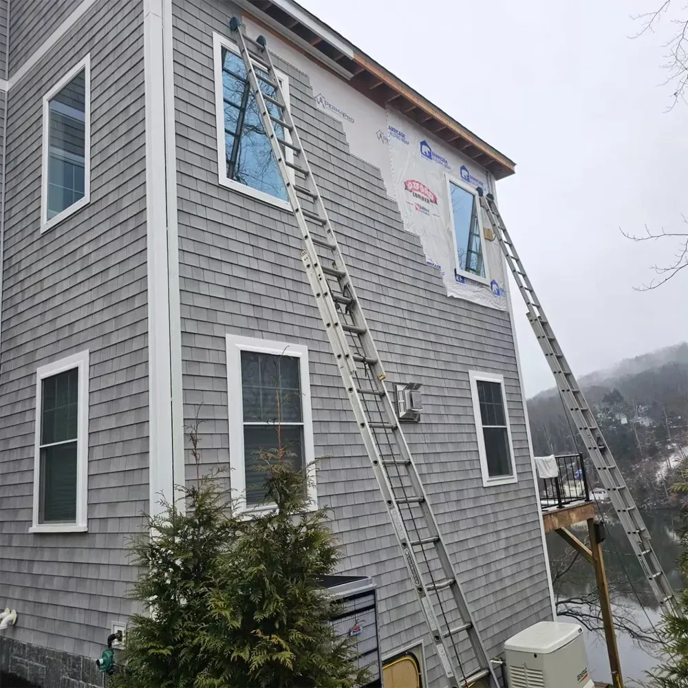 Two extension ladders lean against the gray-shingled exterior of a two-story house undergoing siding repair.