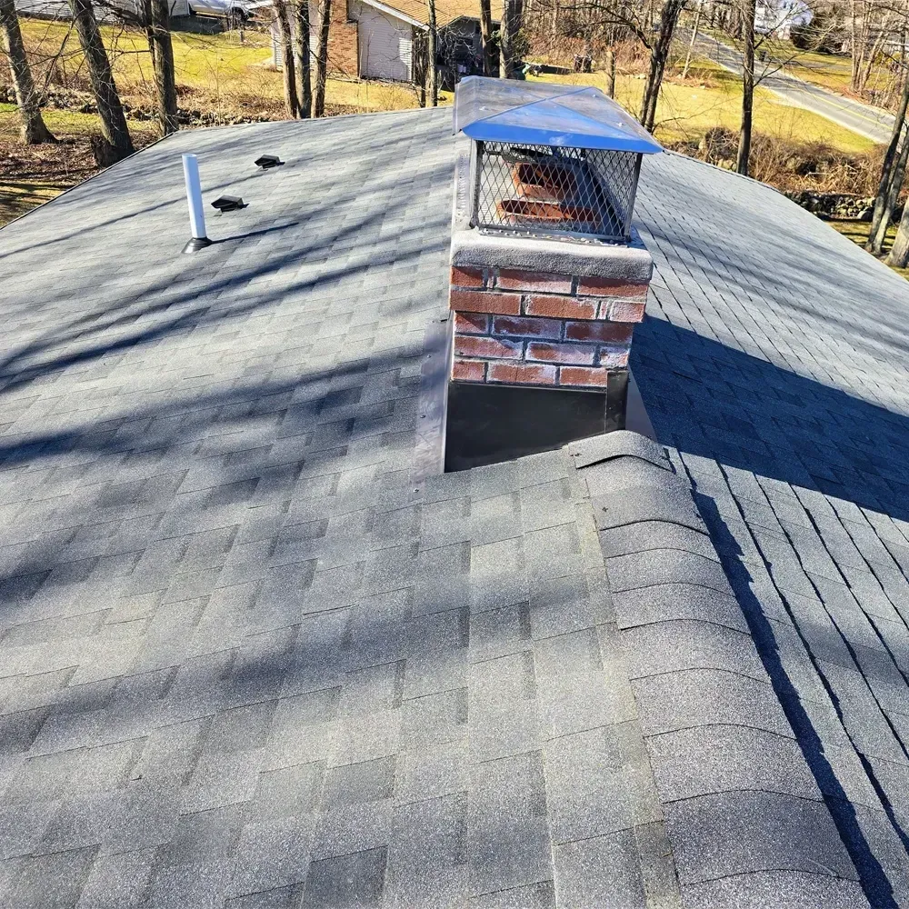 A view from a shingled roof showing a brick chimney with a blue metal cap and a small white pipe vent nearby.