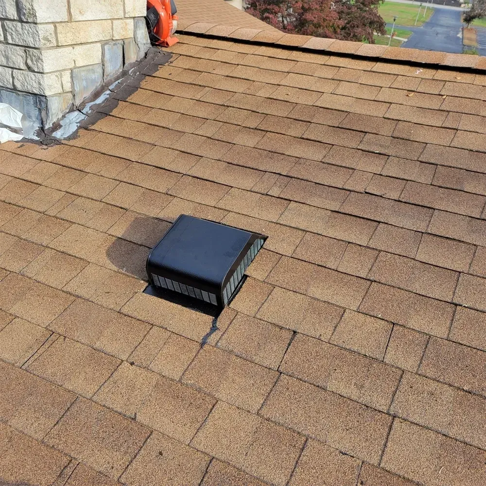 A black square roof vent installed on a brown shingled roof next to a stone chimney.