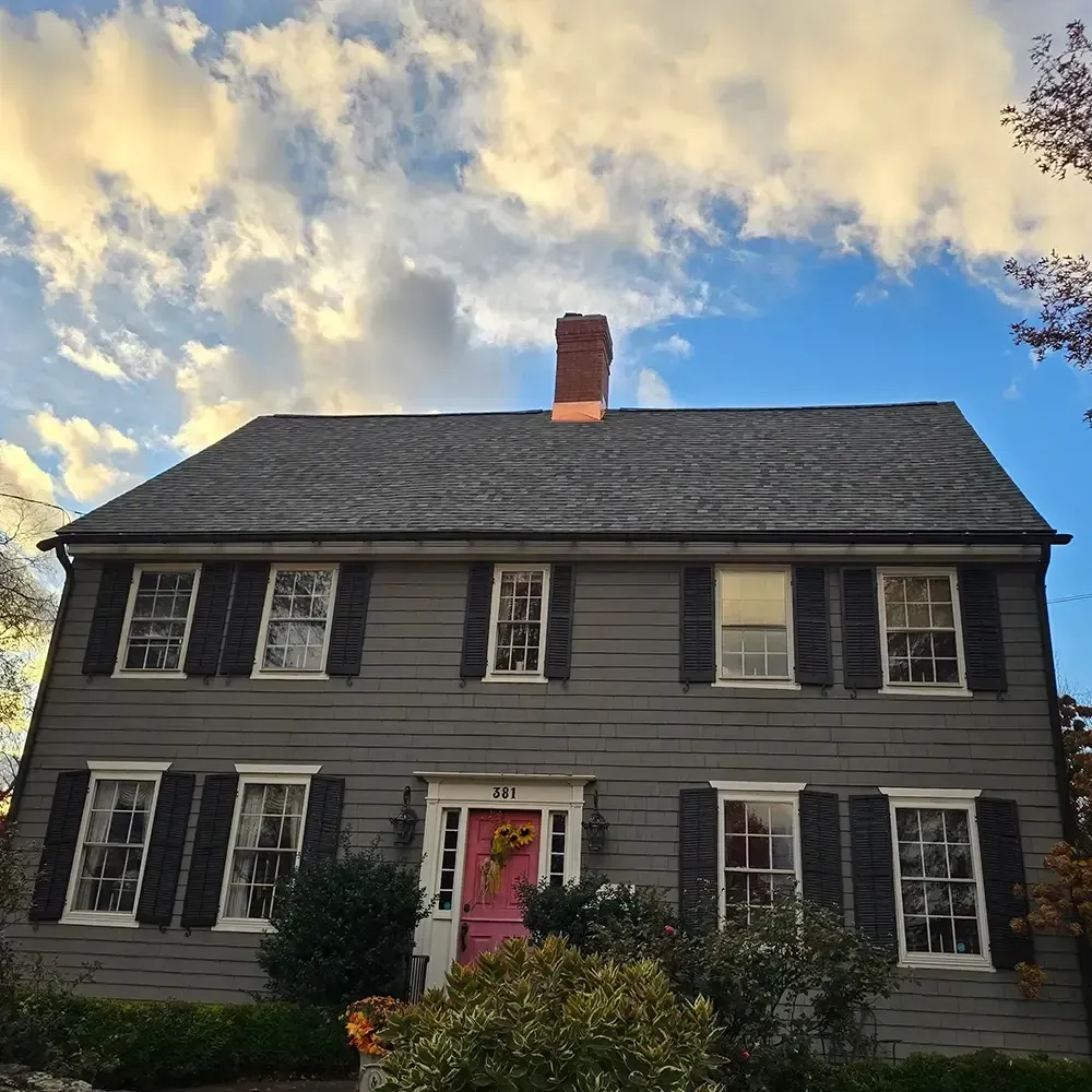 A two-story, gray colonial-style house with black shutters, a bright pink front door, and a chimney against a blue sky.