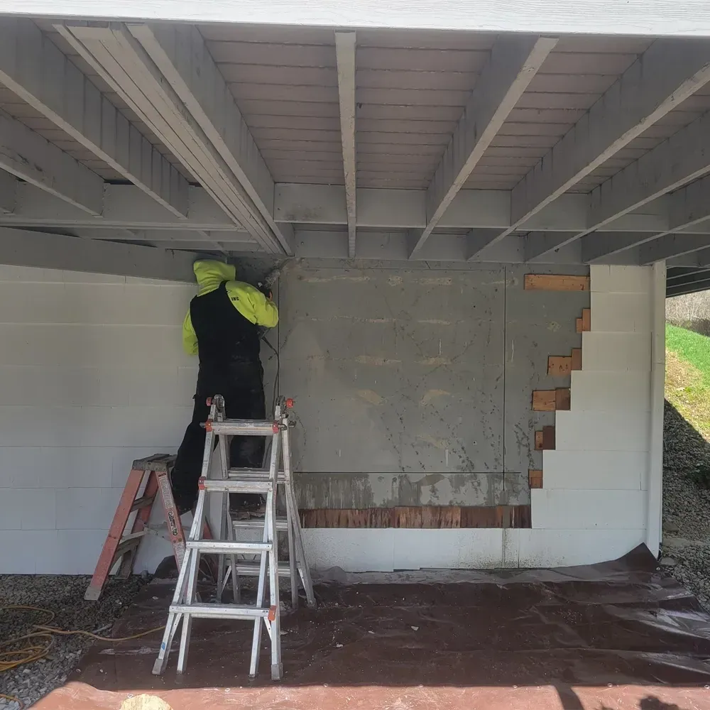 A person in a high-visibility jacket stands on a ladder working on the underside of a deck against a concrete wall.