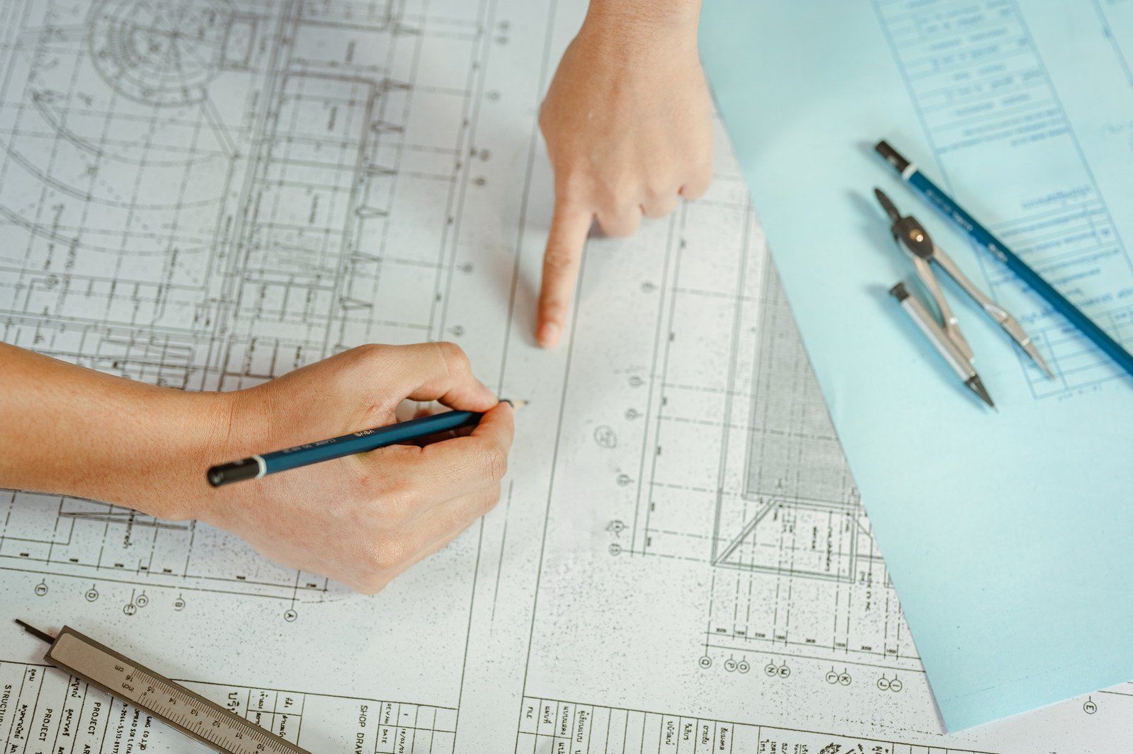 Hands working on blueprints with pencil, compass, and ruler on a desk, discussing design.