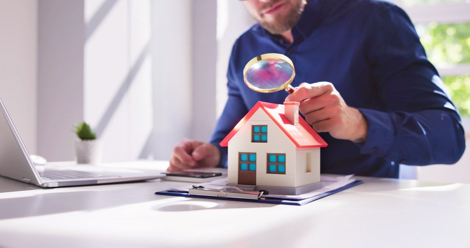 Man inspects a miniature house with a magnifying glass, at a desk with a laptop.