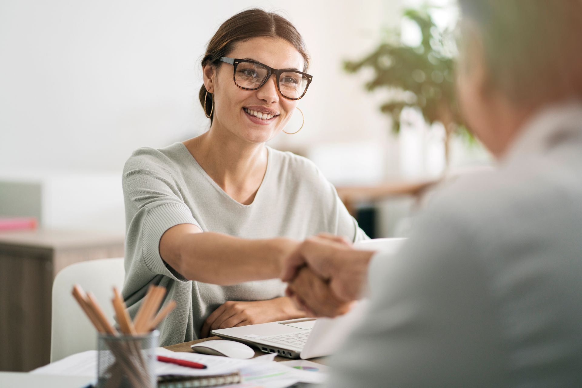 Woman in glasses smiles, shaking hands with a person at a desk in an office setting.