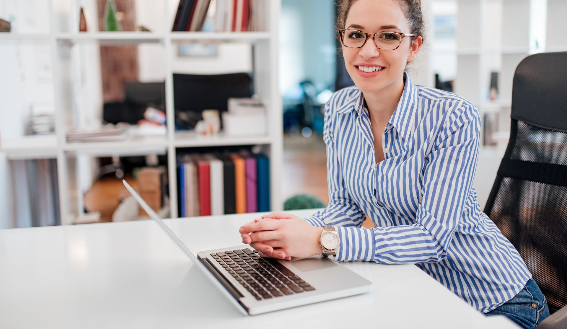 Woman wearing glasses, striped shirt, smiles while sitting at a desk with a laptop in a bright office.