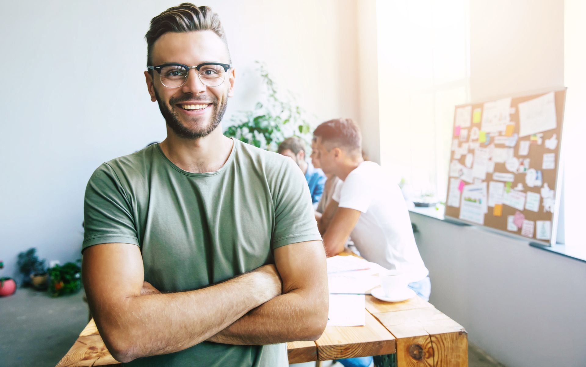 Man with glasses smiles, arms crossed, in office setting; coworkers at table.