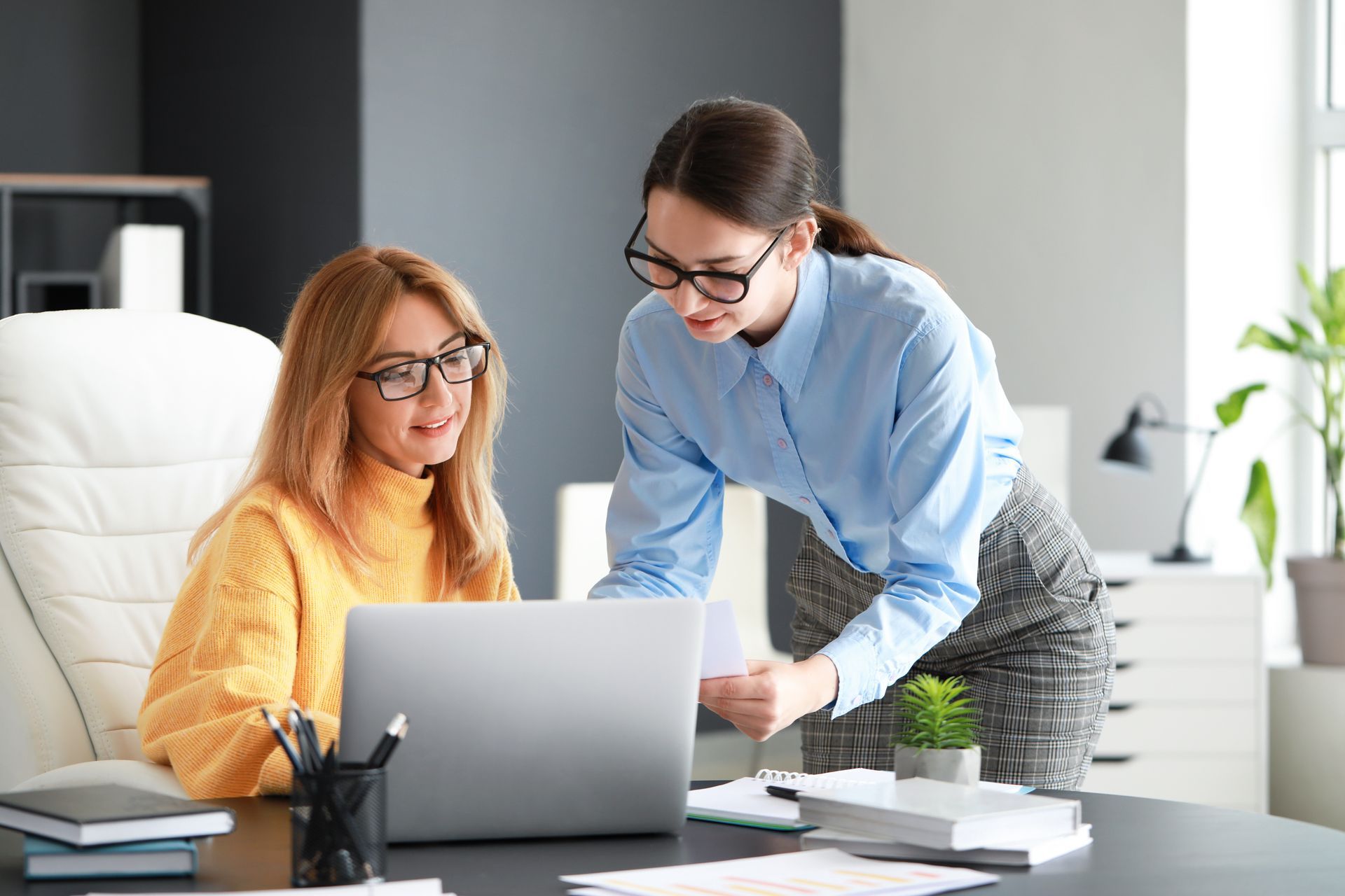 Two women at office desk looking at laptop, one pointing to paper.